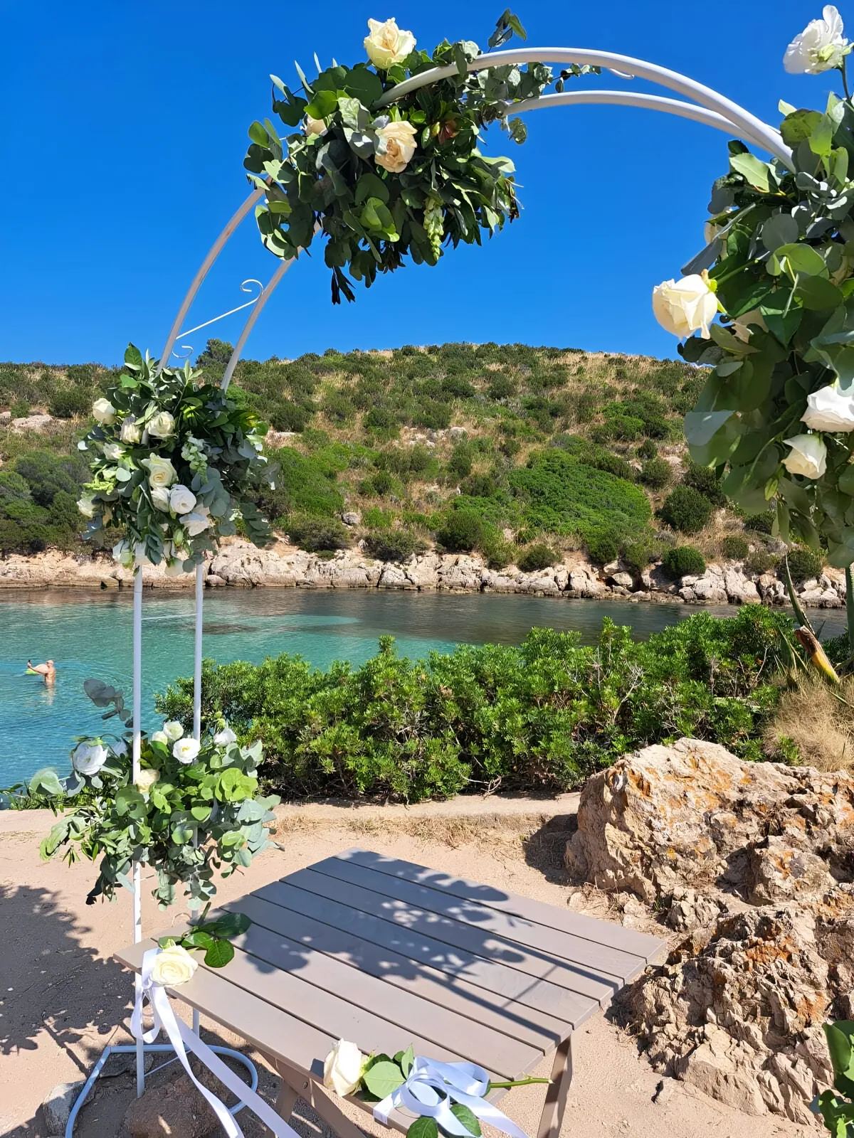 Arco cerimoniale di fiori bianchi tra le rocce, sopra una caletta